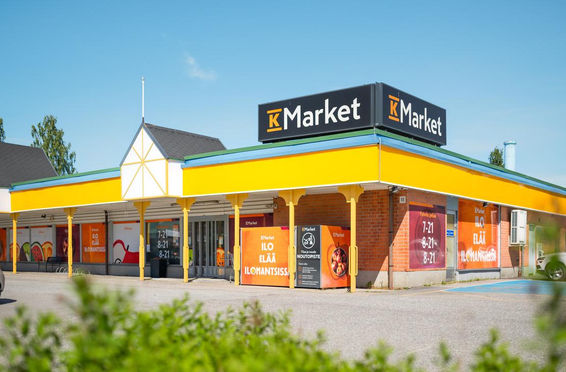 K-Market Ilomantsi -ruokakaupan julkisivu ja sisäänkäynti aurinkoisena päivänä sinisen taivaan alla. Tiilirakennuksen katolla on suuret mustat K-Market-kyltit, ja katon reunaa kiertää keltainen lippa. Ikkunoissa ja seinissä on oransseja opasteita, joissa lukee ”ILO ELÄÄ ILOMANTSISSA” sekä kaupan aukioloajat. Sisäänkäynnin lähellä on noutopistekaappi, ja etualalla kasvaa vihreää pensaikkoa.