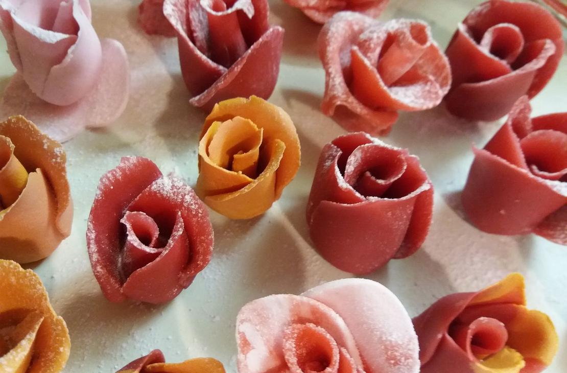 Lähikuva useista syötävistä ruusunmuotoisista koristeista tai makeisista, jotka on tehty sokerimassasta tai marsipaanista, mahdollisesti Kotipari Lämmin Halaus Oy:n tuotteita. Ruusut ovat eri sävyisiä, mukaan lukien vaaleanpunainen, koralli ja oranssi, ja ne on aseteltu vaalealle pinnalle.