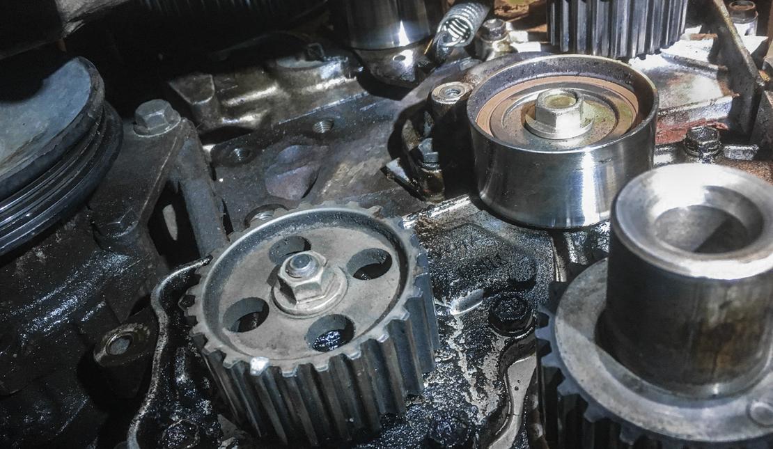 Lähikuva autonmoottorin osista, todennäköisesti jakopään alueelta. Kuvassa näkyy kaksi hammaspyörää ja yksi kiristinrulla tai vastaava metallinen pyörä. Osat ovat tummat, kiiltävät ja mahdollisesti öljyiset. Kohtaus on otettu moottoritilassa korjauksen tai huollon aikana.