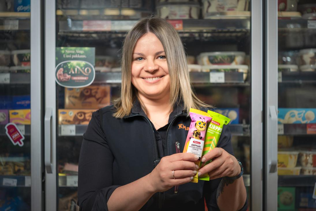 Mustaan K-Market-paitaan pukeutunut työntekijä hymyilee ja pitelee käsissään Pirkka Amppari- ja Pirkka Kirppu -mehujäitä K-Market Ilomantsissa. Hänen takanaan on lasiovinen pakastekaappi, jonka hyllyillä on erilaisia jäätelöitä ja jonka ovessa on pyöreä Aino-jäätelön tarra.