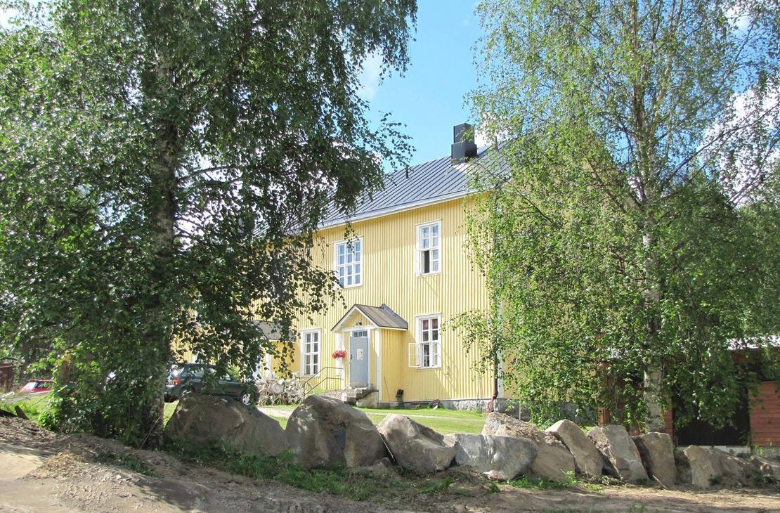 Katri Vala -kulttuurikeskuksen keltainen, kaksikerroksinen puurakennus aurinkoisena kesäpäivänä vihreiden koivujen ympäröimänä. Etualalla on hiekkamaata ja suurista luonnonkivistä koostuva matala muuri. Rakennuksessa on tumma harjakatto, valkoiset ikkunanpuitteet ja pieni katettu kuisti harmaan ulko-oven päällä.