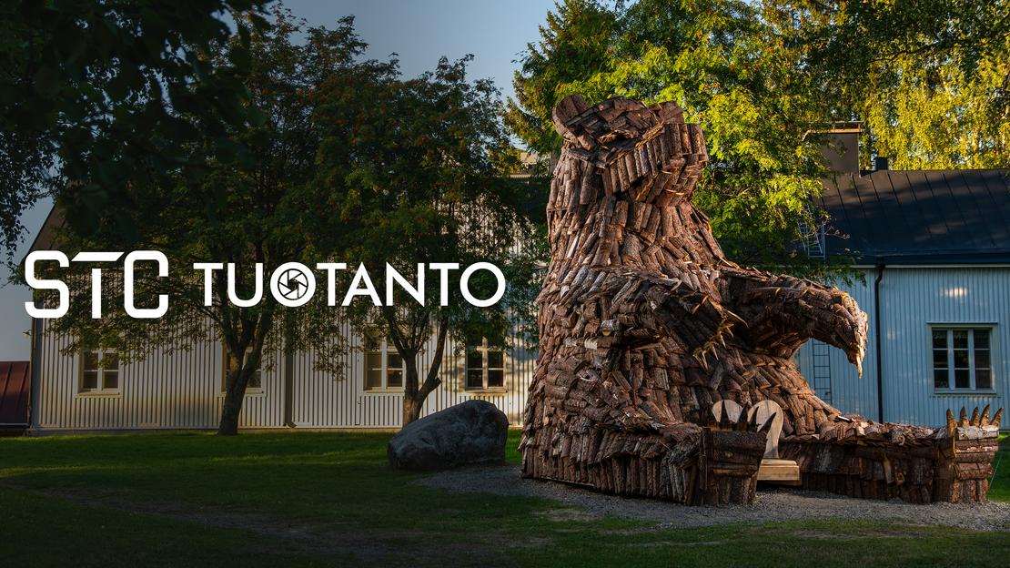 Kuva esittää pihanurmea, jolla istuu suuri, karhunmuotoinen veistos tai patsas, joka on tehty risuista, puunpaloista tai muusta tummasta materiaalista. Patsas on sijoitettu historiallisen, vaalean puurakennuksen (oikealla) ja valkoisen rakennuksen (vasemmalla) eteen. Kuvan päällä lukee valkoisella, läpikuultavalla tekstillä "STC TUOTANTO", ja T-kirjainten välissä on pieni pyöreä logo.