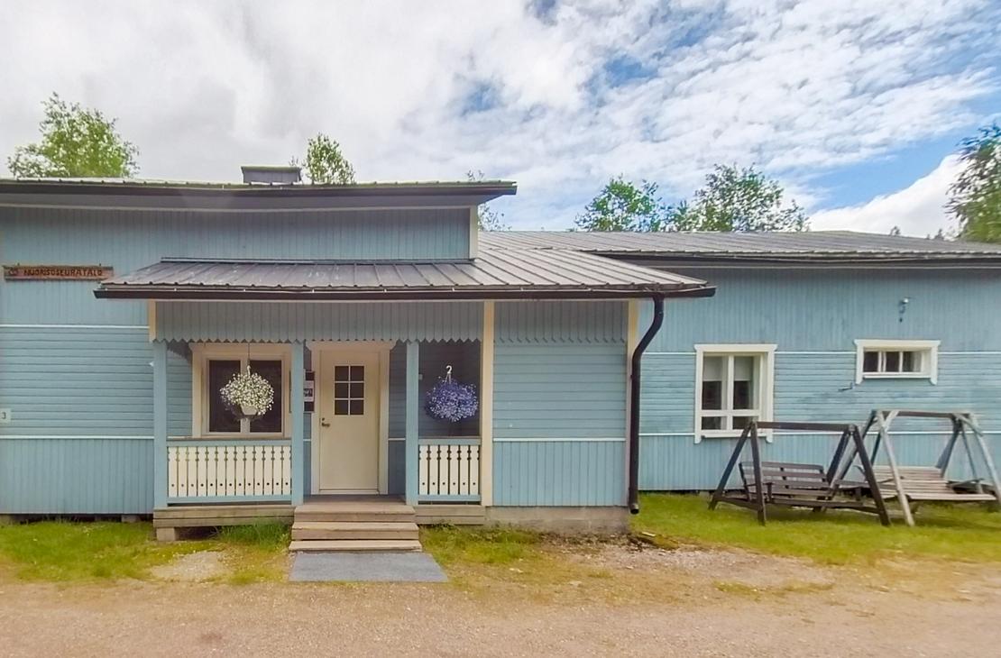 Kuva esittää matalaa, modernia, vaaleansinistä puutaloa tai rakennusta, jossa on tumma katto. Talon keskellä on pieni, katettu kuisti, jonka edessä on portaat. Kuistin oven yläpuolella näkyy pyöreä, koristeellinen valaisin tai symboli. Talon edessä on soraa tai hiekkamaata, ja oikealla sivulla on tumma, metallinen keinu tai teline nurmikolla. Taustalla näkyy rehevää kasvillisuutta ja metsää.