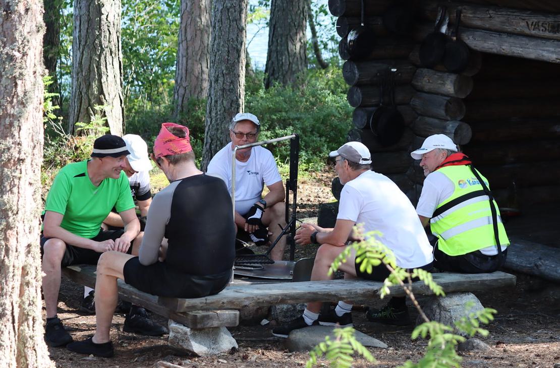 Ryhmä miehiä istuu puupenkeillä nuotiopaikan äärellä metsässä sijaitsevan hirsilaavun edustalla.