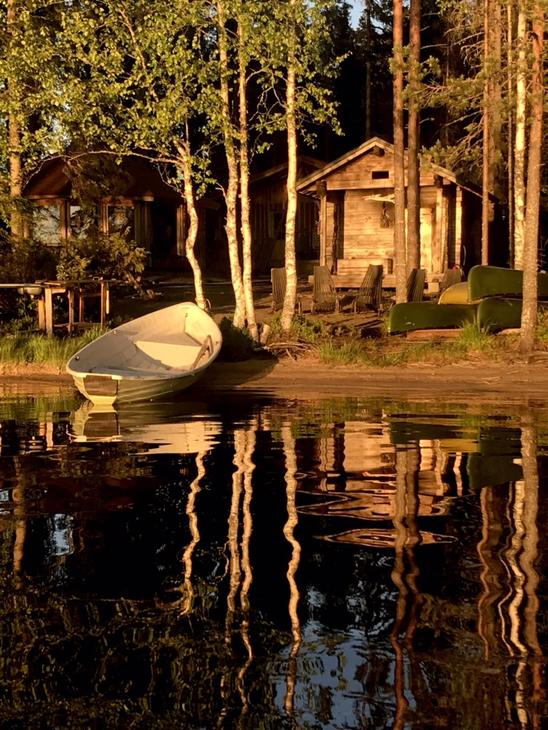 Kuva esittää Kotakylän rantasaunaa tai -mökkialuetta auringonlaskun aikaan. Etualalla on tummaa, tyyntä vettä, johon rannan puut ja rakennukset heijastuvat pitkinä varjoina. Rannalla, hiekalla tai nurmella, seisoo vaalea soutuvene kallellaan. Taustalla pystyssä olevien puunrunkojen takana näkyy kaksi pientä puurakennusta (mökit tai aitat), joita valaisee matala, lämmin auringonvalo.