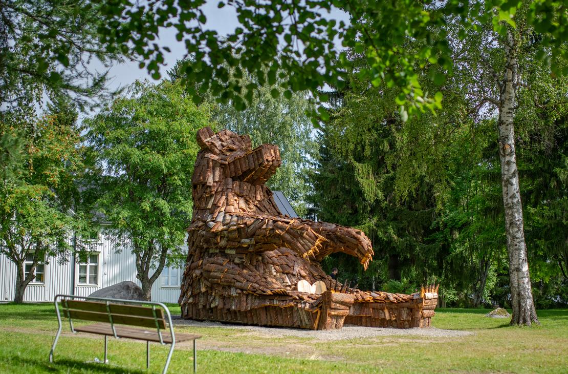L&auml;hikuva suuresta, puusta tehdyst&auml; karhupatsaasta, ILOKARHU -nimisest&auml; veistoksesta Ilomantsista. Karhu on koottu useista pienemmist&auml; puupaloista ja laudoista. Kuvassa n&auml;kyy selv&auml;sti karhun karkea pintakuvio. Taustalla on vihre&auml;&auml; kasvillisuutta.