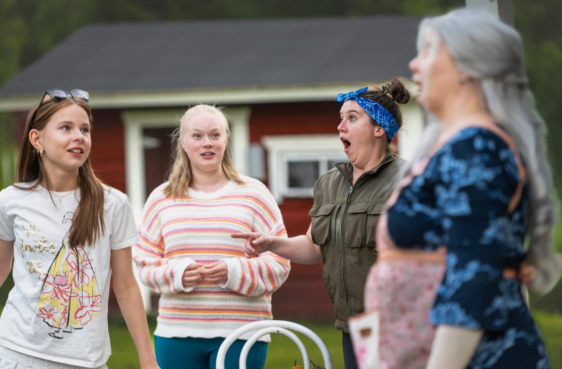 Möhkön kesäteatterin Peitetarina-näytelmän kohtaus, jossa neljä naisnäyttelijää esiintyy ulkonäyttämöllä punaisen rakennuksen edustalla. Oikealla etualalla näkyy epätarkkana sivuprofiili harmaatukkaisesta naisesta. Keskellä sinipäähineinen nainen osoittaa vasemmalle suu ammollaan hämmästyksestä, ja vasemmalla kaksi naista seuraa tilannetta ilmeikkäästi.