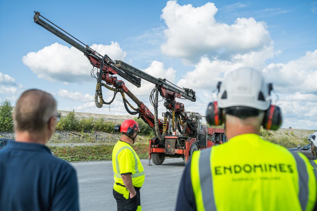 Kolme henkilöä seisoo selin kameraan ja katsoo suurta punaista kaivosporauslaitetta, jonka pitkä puomi on suunnattu viistosti kohti taivasta. Etualalla oikealla olevalla henkilöllä on päässään valkoinen suojakypärä ja yllään keltainen huomioliivi, jonka selässä lukee "ENDOMINES". Tapahtuma sijoittuu sorakentälle aurinkoisena päivänä, ja taustalla on sininen taivas valkoisilla pilvillä.