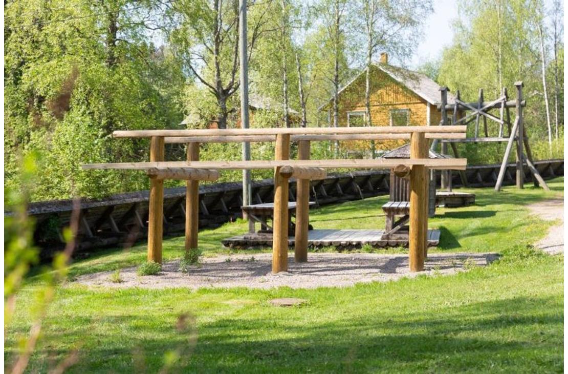 Kuva esittää Herralankosken puiston matonpesupaikan yhteydessä sijaitsevaa puista matonkuivaustelinettä. Kaksitasoinen teline seisoo hiekkapohjalla vihreällä nurmikentällä. Telineen takana näkyy vanha, keltainen puutalo ja puita, jotka ovat täydessä kesävihreydessä.