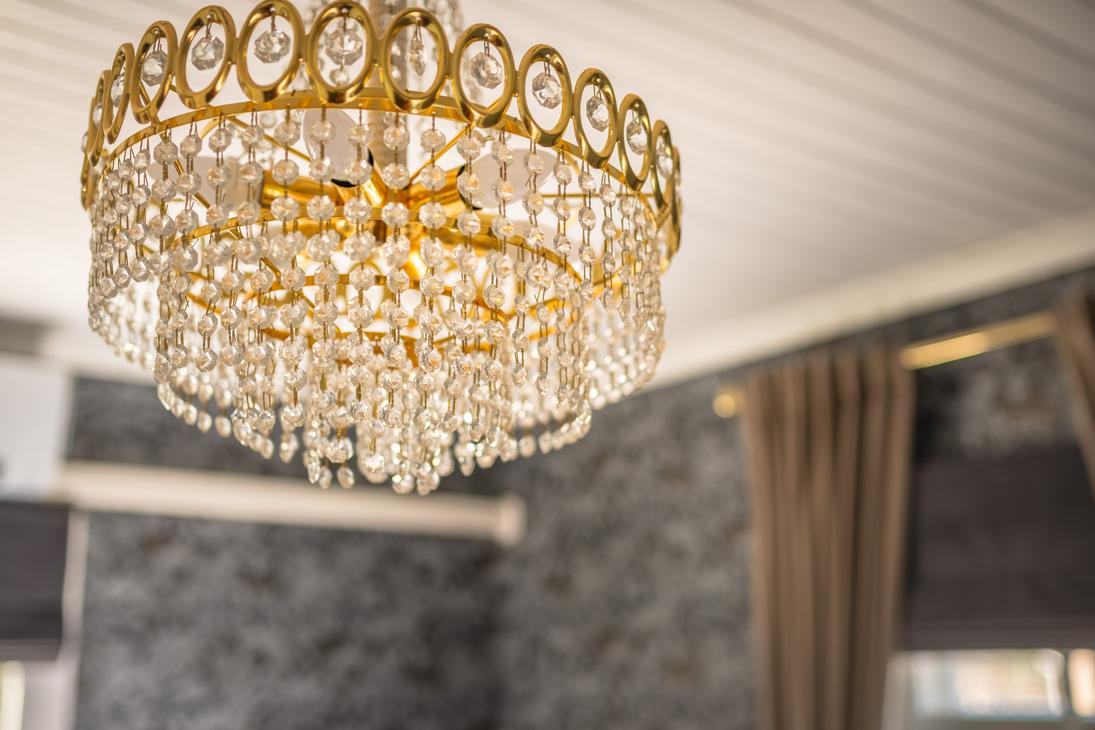 Lähikuva kristallikruunusta Matkailukeskus Puu-Juuka Oy:n sisätiloissa. Kruunu on suuri, kultaisen värinen ja siinä on useita kerroksia roikkuvia kristalleja. Kruunu roikkuu valkoisesta, lankulla vuoratusta katosta. Tausta on epätarkka, mutta siellä näkyy tummanharmaata tai mustaa seinämateriaalia ja ruskeita verhoja.