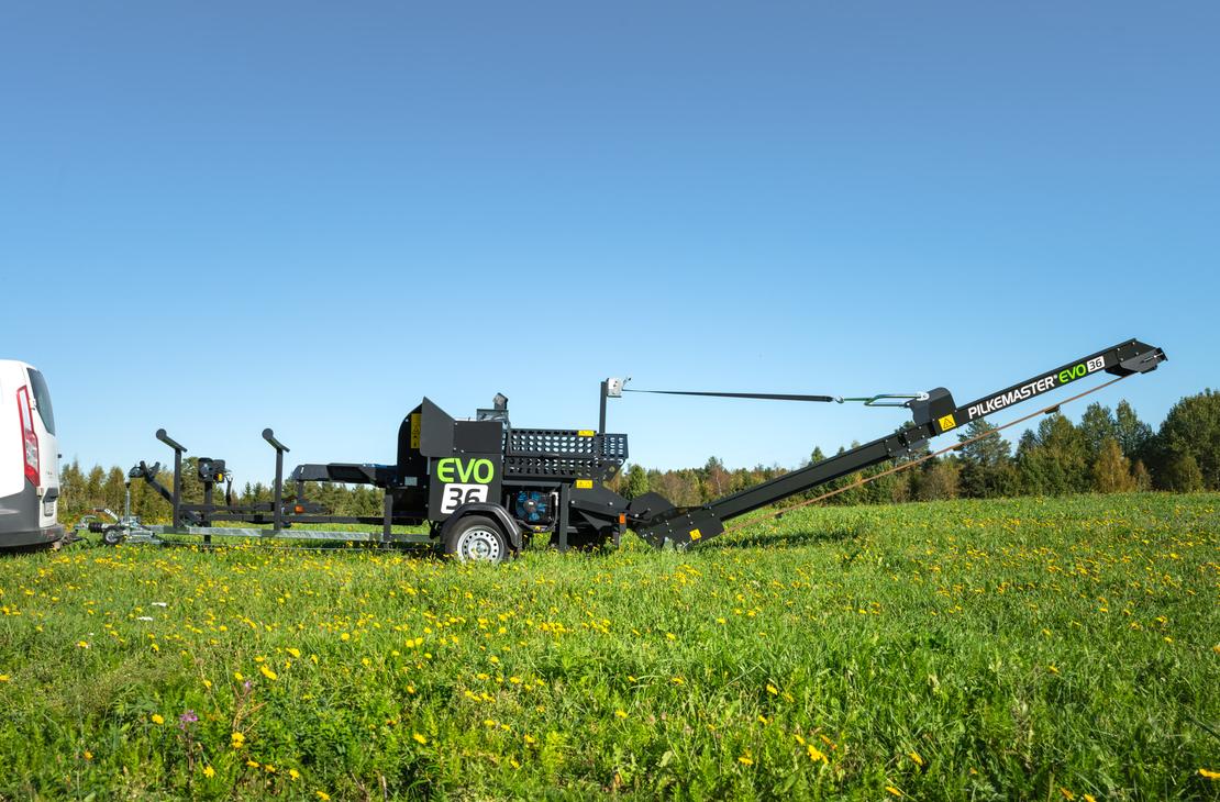 KLAPikosen musta ja vihreä Pilkemaster EVO 36 -klapikone seisoo aurinkoisella, keltaisten voikukkien täplittämällä niityllä valkoisen pakettiauton perässä, kirkkaansinisen taivaan alla.