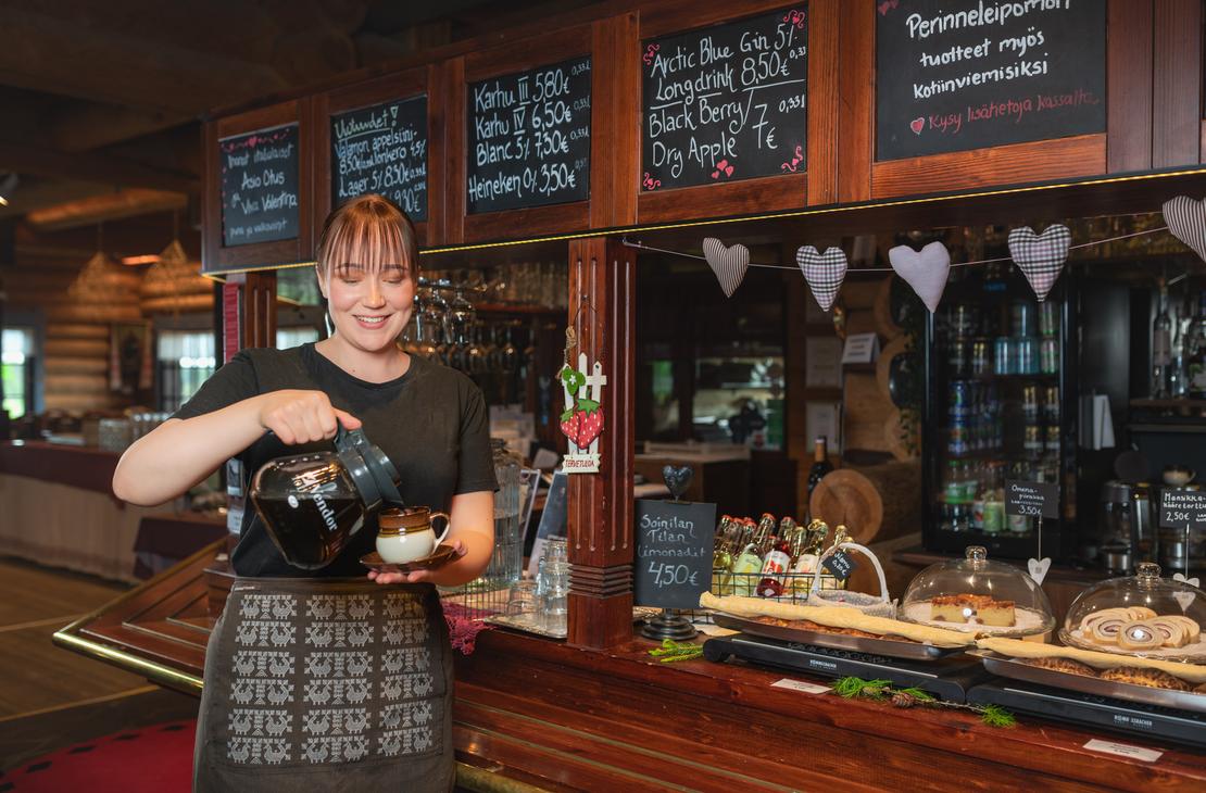 Hymyilevä, karjalaiseen essuun pukeutunut tarjoilija kaataa kahvia pannusta kuppiin Ravintola Parppeinpirtin tiskillä Ilomantsissa. Taustalla näkyy hirsiseinä, hinnastoliitutauluja ja vitriinissä olevia leivonnaisia.