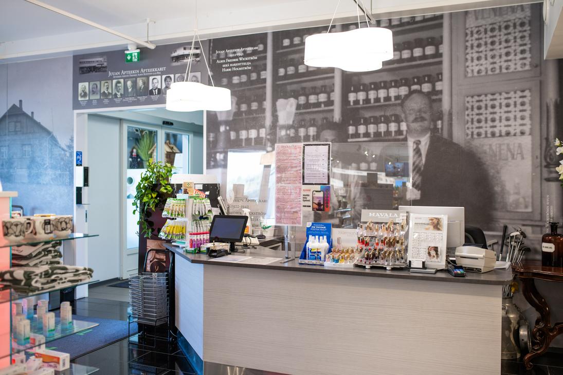Kuva esittää Juuan Apteekin sisätilaa ja palvelutiskiä. Etualalla on harmaa palvelutiski, jonka takana on seinällä mustavalkoinen, suuri valokuva vanhanaikaisesta apteekista tai apteekkarista hyllyineen. Tiskillä on erilaisia apteekkituotteita. Tila on hyvin valaistu.