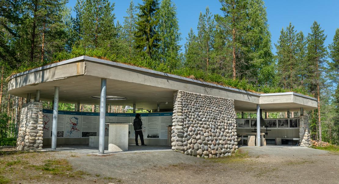 Kuva Öykkösenvaaran taistelualueen taukopaikasta tai museorakennuksesta. Se on moderni, matala, osittain avoin katos, jossa on vihreä, maanpeittämä katto. Rakenne koostuu betonista ja pyöreillä luonnonkivillä verhoilluista pylväistä. Rakennuksen alla on taukopaikka tai näyttelytila. Rakennus sijaitsee metsän reunassa soraisella maapohjalla.