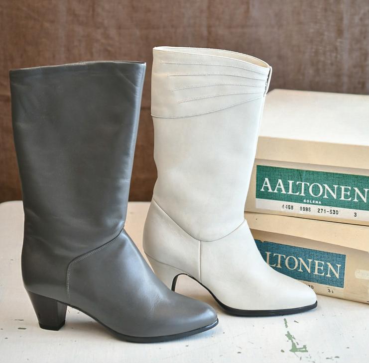 Harmaa ja valkoinen siro nahkasaapas aseteltuna vierekkäin. Taustalla näkyy Aaltonen-kenkälaatikoita.