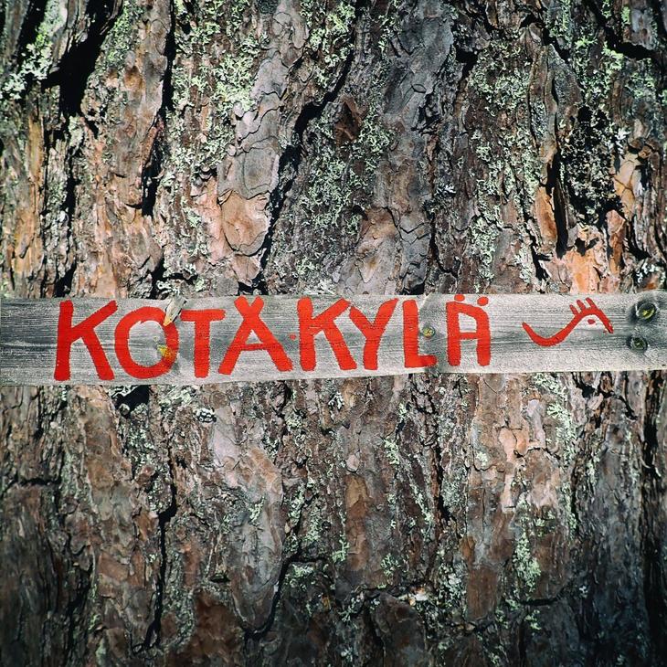 Lähikuva vanhan puun (todennäköisesti männyn) karkeasta, halkeilevasta kuoresta. Puunrunkoa vasten on kiinnitetty vaakasuora, vaaleampi puulauta, johon on maalattu kirkkaan punaisella tekstillä sana "KOTAKYLÄ". Tekstin oikeassa reunassa on pieni, punainen, tyylitelty kuvio, joka muistuttaa sarvipäätä tai eläintä.