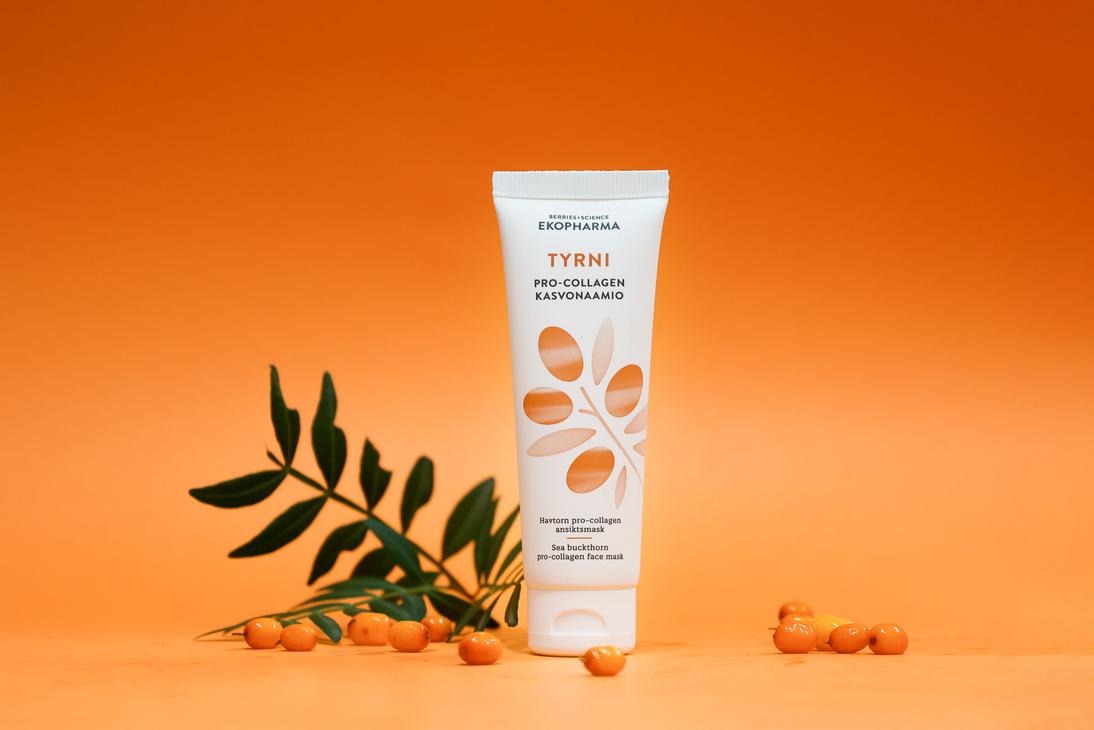 Ekopharma Tyrni Pro-Collagen -kasvonaamiotuubi oranssilla taustalla. Tuubin vieressä on vihreä tyrninoksa ja muutama tyrnimarja oranssilla alustalla.