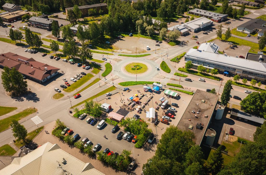 Korkealta otettu ilmakuva Ilomantsin keskustan liikenneympyr&auml;st&auml; ja sen vieress&auml; sijaitsevasta torialueesta, jossa on v&auml;rikk&auml;it&auml; myyntitelttoja, ihmisi&auml; ja ymp&auml;rill&auml; runsaasti pys&auml;k&ouml;ityj&auml; autoja aurinkoisena kes&auml;p&auml;iv&auml;n&auml;.