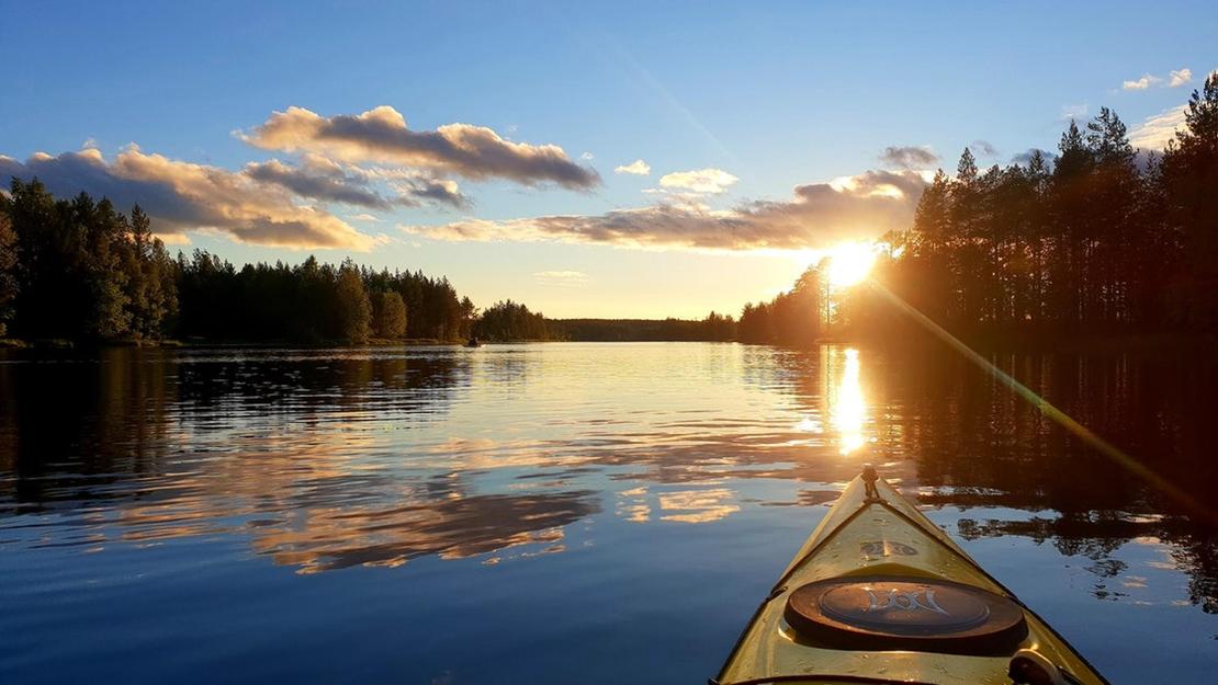 Kuva on otettu kajakista tai kanootista, ja se esittää tyyntä järveä auringonlaskun aikaan. Kajakin keula (vaaleankeltainen) on etualalla. Aurinko laskee tai nousee suoraan edessä, luoden kirkkaan heijastuksen veteen, joka on sinistä ja heijastaa taivaan pilviä. Järveä reunustaa tumma, tiheä metsä molemmin puolin. Kuva liittyy todennäköisesti Äksyt Ämmät -yrityksen melontapalveluihin.