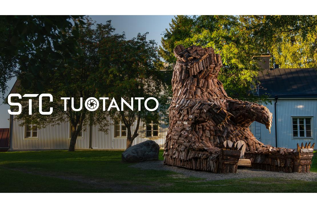 Kuva esitt&auml;&auml; pihanurmea, jolla istuu suuri, karhunmuotoinen veistos tai patsas, joka on tehty risuista, puunpaloista tai muusta tummasta materiaalista. Patsas on sijoitettu historiallisen, vaalean puurakennuksen (oikealla) ja valkoisen rakennuksen (vasemmalla) eteen. Kuvan p&auml;&auml;ll&auml; lukee valkoisella, l&auml;pikuultavalla tekstill&auml; "STC TUOTANTO", ja T-kirjainten v&auml;liss&auml; on pieni py&ouml;re&auml; logo.