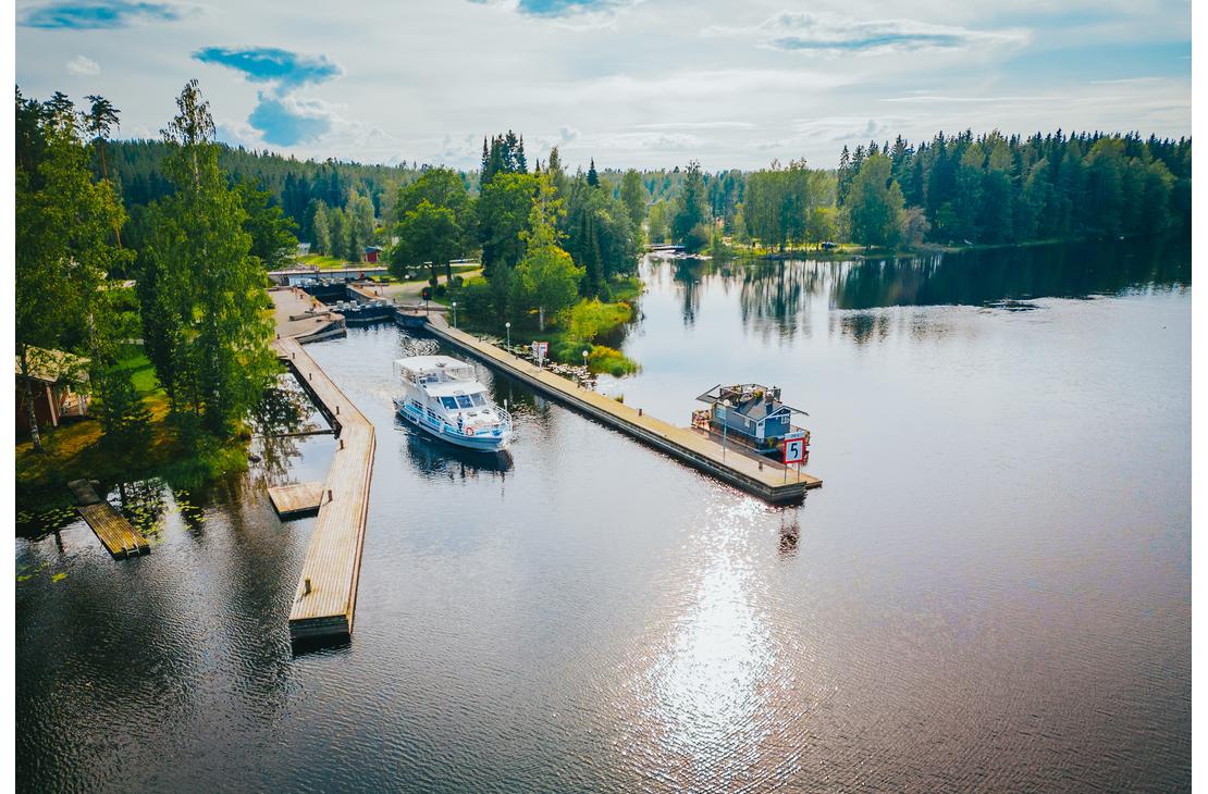 Ilmakuva tyynestä vesistöstä aurinkoisena päivänä, joka liittyy Blue Lake Cruisesin toiminta-alueeseen. Kuvassa näkyy kaksi venelaituria ja niihin kiinnitettynä useita aluksia, mukaan lukien suurempi, valkoinen risteilyalus ja pienempi vene. Laitureiden takana näkyy osa maata, mahdollisesti kanava tai sulku (vasemmalla), ja aluetta ympäröi tiheä, vehreä metsä. Auringonvalo heijastuu kirkkaasti veden pintaan oikealla.