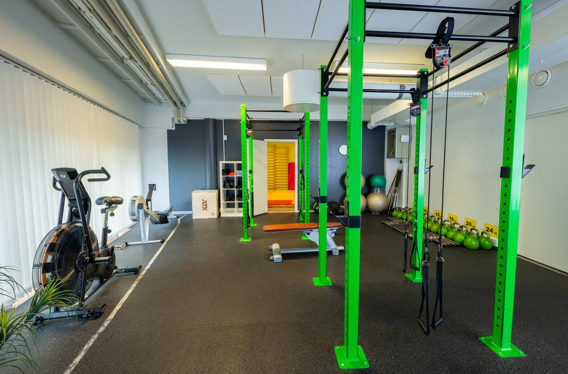 Kuva esittää Kinesiocenter Kuntoilukeskuksen harjoitustilaa. Lattiana on tumma kuminen lattia, ja tilassa on kirkkaan limevihreitä metallisia telineitä crossfit-tyyliseen harjoitteluun. Vasemmalla näkyy kuntopyörä tai ergometri. Oikealla taustalla on rivi kahvakuulia ja keskellä näkyy avoimen oven kautta seuraava huone, joka on keltainen ja punainen.