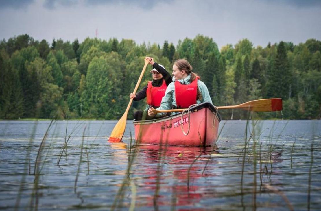 Kaksi henkilöä, nainen etualalla ja mies takana, melovat punaisella avokanootilla tyynellä järvellä. Molemmilla on yllään kirkkaan punaiset pelastusliivit. He pitelevät puisia meloja, joissa on keltaiset lavat. Etualalla vedestä nousee pitkiä, ohuita ruohoja. Taustalla on tiheä, vihreä metsä. Kuva liittyy todennäköisesti Äksyt Ämmät -yrityksen melontapalveluihin.