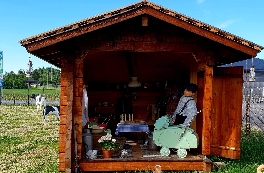 Kuva esitt&auml;&auml; Juuan Apteekin perinnepihaa aurinkoisena p&auml;iv&auml;n&auml;. Etualalla on pieni, puinen katos tai koju tummasta puusta, jossa on n&auml;ytteill&auml; vanhanaikaisia esineit&auml;, kuten nukke tai patsas perinteisiss&auml; vaatteissa keinutuolissa sek&auml; p&ouml;yt&auml;liinoja ja kukkia. Vasemmalla nurmialueella n&auml;kyy ihmisi&auml; ja maalattuja lehm&auml;patsaita tai koristeita. Taustalla erottuu lipputanko ja kadunvarsirakennuksia.