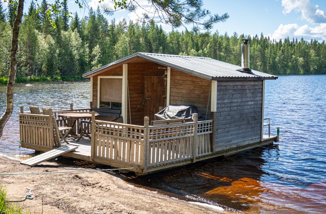 Puurakenteinen saunalautta ankkuroituna hiekkaiselle rannalle tyynen järven äärellä. Lautalla on terassi, jossa on pöytäryhmä ja grilli, sekä korkea metallinen piippu.