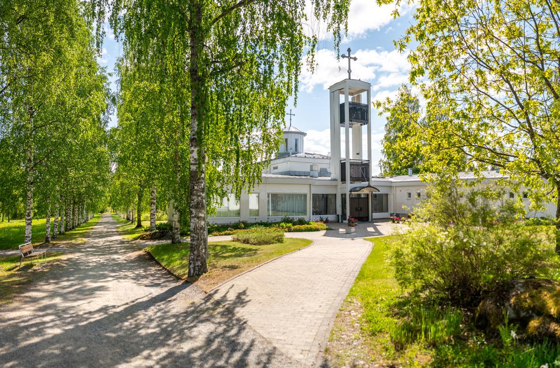 Laajakulmakuva Lintulan luostarin sisäänkäynnille aurinkoisena päivänä. Vasemmalla reunustaa vehreä koivukuja, ja oikealla laatoitettu käytävä johtaa moderniin, valkoiseen luostarirakennukseen, jossa on erillinen kellotapuli ja risti.