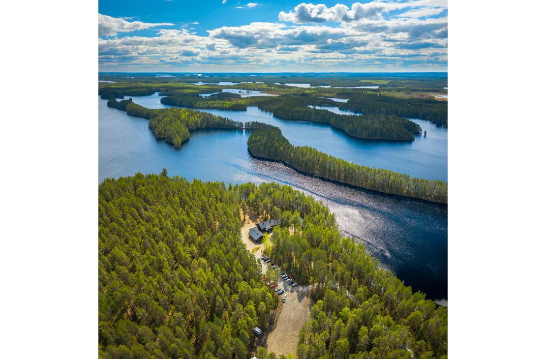 Ilmakuva Petkelj&auml;rvi Centerin alueesta ja Petkelj&auml;rven kansallispuiston maisemista. Etualalla on tihe&auml;&auml;, vihre&auml;&auml; mets&auml;&auml; ja avoin, hiekkainen alue, jossa n&auml;kyy muutama tummakattoinen rakennus (Petkelj&auml;rvi Center). Taustalla avautuu laaja j&auml;rvimaisema, jossa tummansininen vesi kietoutuu lukuisten vihreiden harjujen ja niemien ymp&auml;rille. Kirkas sininen taivas, jossa on valkoisia pilvi&auml;.