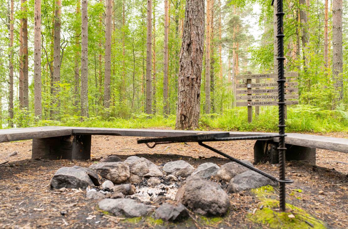 Lähikuva maastossa olevasta nuotiopaikasta ja grilliritilästä. Taustalla näkyy puinen opasteviitta, jossa on reittien ja lampien nimiä, kuten Hiislampi ja Taitajan taival.