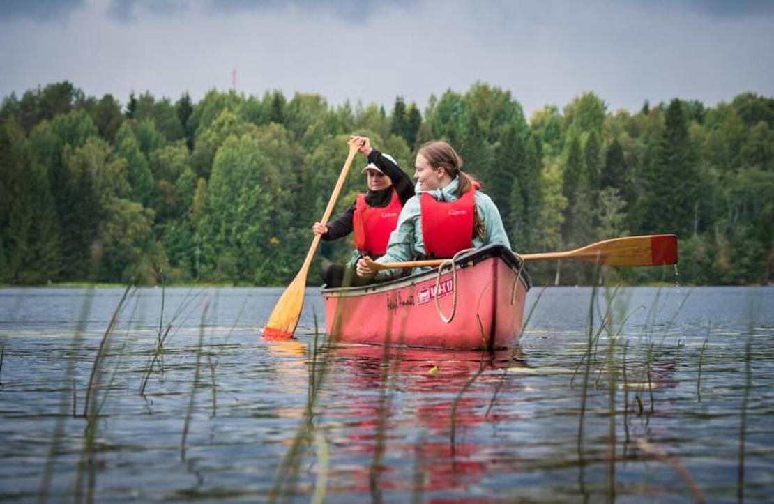 Kaksi henkilöä, nainen etualalla ja mies takana, melovat punaisella avokanootilla tyynellä järvellä. Molemmilla on yllään kirkkaan punaiset pelastusliivit. He pitelevät puisia meloja, joissa on keltaiset lavat. Etualalla vedestä nousee pitkiä, ohuita ruohoja. Taustalla on tiheä, vihreä metsä. Kuva liittyy todennäköisesti Äksyt Ämmät -yrityksen melontapalveluihin.