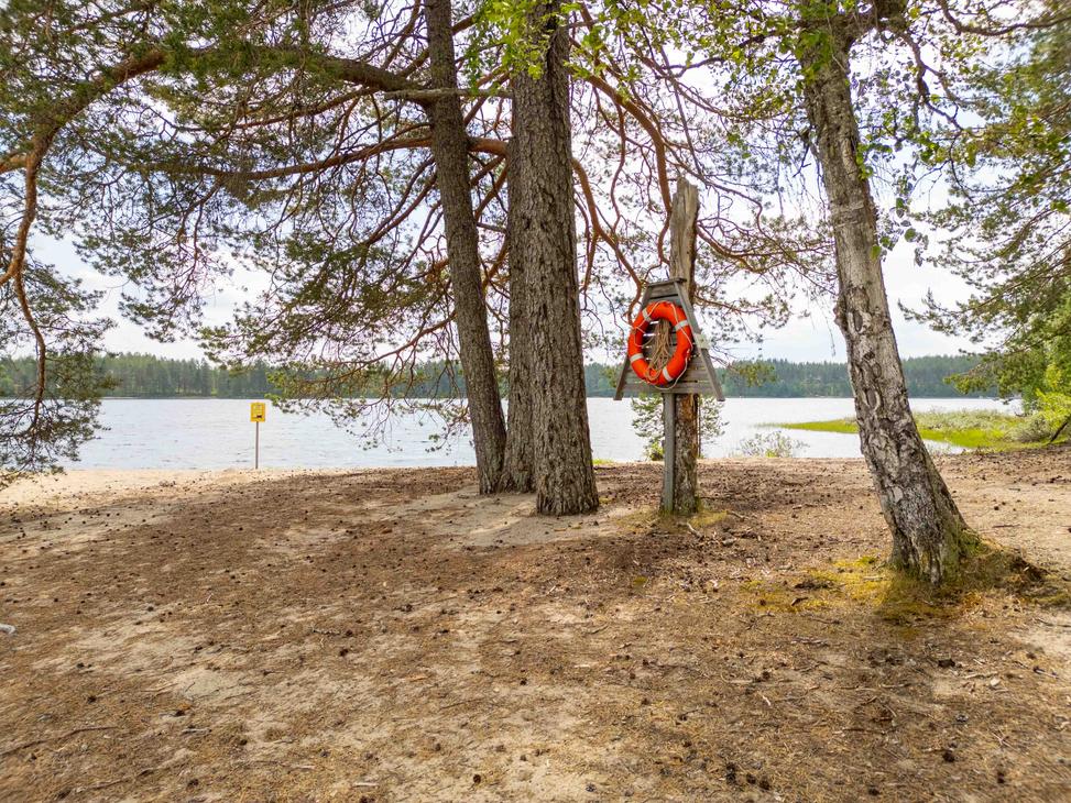 Punavalkoinen pelastusrengas kiinnitettynä puiseen telineeseen rantamännikön siimeksessä. Taustalla avautuu vaalea hiekkaranta ja tyyni järvimaisema.
