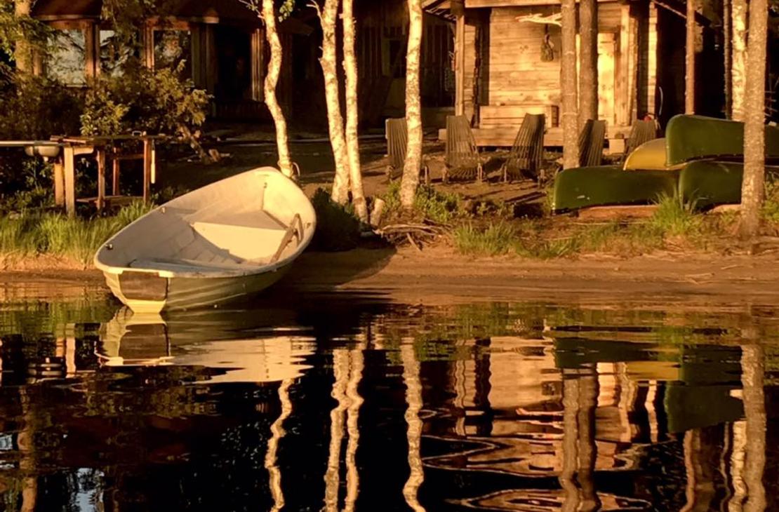 Kuva esittää Kotakylän rantasaunaa tai -mökkialuetta auringonlaskun aikaan. Etualalla on tummaa, tyyntä vettä, johon rannan puut ja rakennukset heijastuvat pitkinä varjoina. Rannalla, hiekalla tai nurmella, seisoo vaalea soutuvene kallellaan. Taustalla pystyssä olevien puunrunkojen takana näkyy kaksi pientä puurakennusta (mökit tai aitat), joita valaisee matala, lämmin auringonvalo.