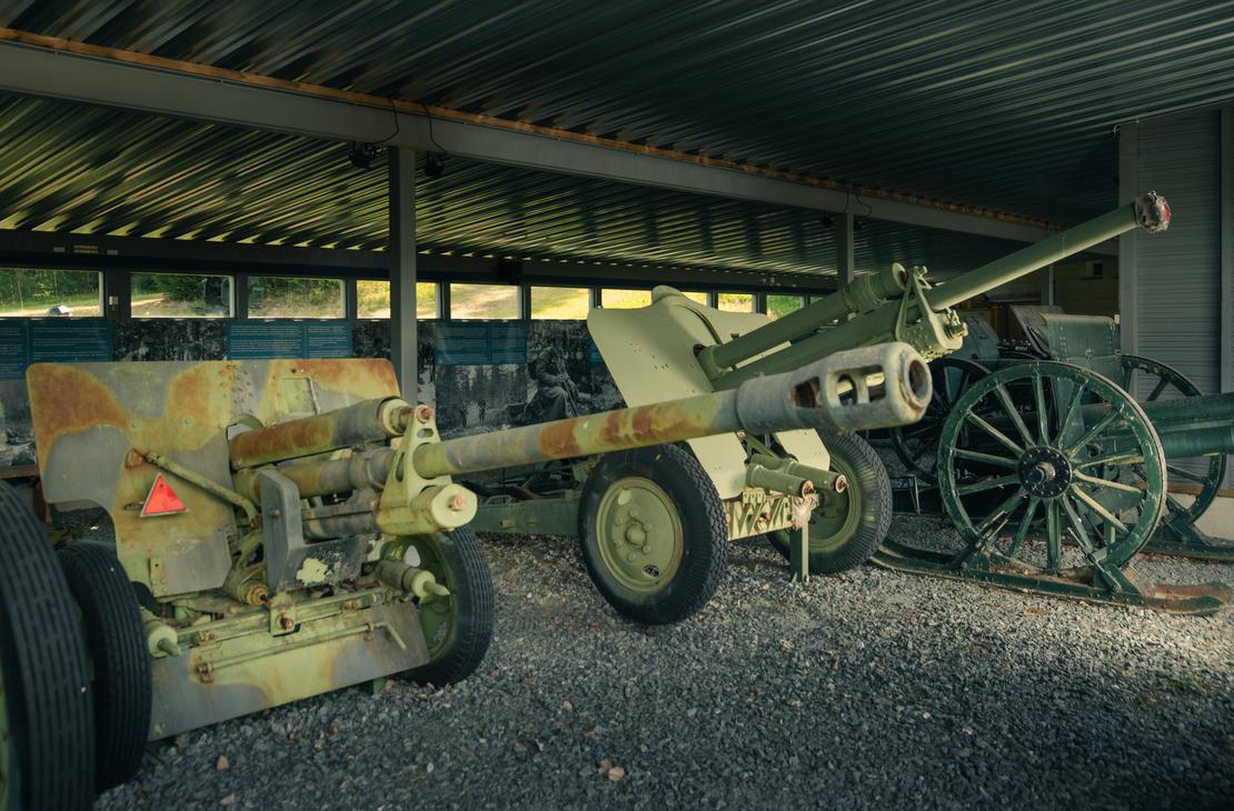 Kuva Taistelijan talon tykkikatoksesta esittää rivin historiallisia tykkejä ja aseita suojassa. Etualalla on harmaa-vihreä, maastokuvioitu panssarintorjuntatykki pyörillä. Sen takana on toinen, samankaltainen tykki, ja oikealla näkyy vanhempi, vihreä kenttätykki suurilla puupyörillä. Katoksen lattia on soraa tai hiekkaa, ja tausta on tumma.