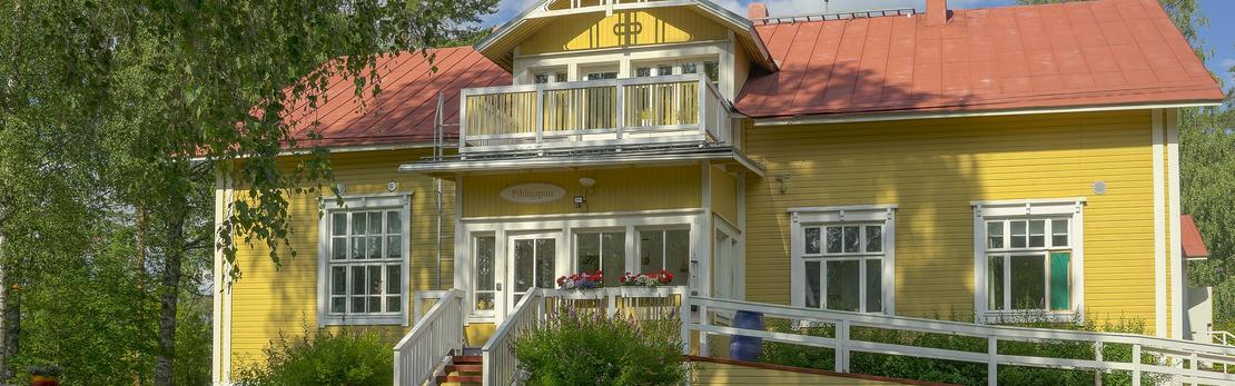 Lähikuva Äksyt Ämmät -yrityksen rakennuksesta, joka on keltainen, perinteinen puutalo punaisella harjakatolla. Rakennuksessa on toisessa kerroksessa pieni, valkokaiteinen parveke ja alakerrassa valkoiset ikkunankarmit. Kuvan edessä näkyy valkoista aitaa tai ramppia. Rakennusta reunustaa vasemmalla vihreät puut.