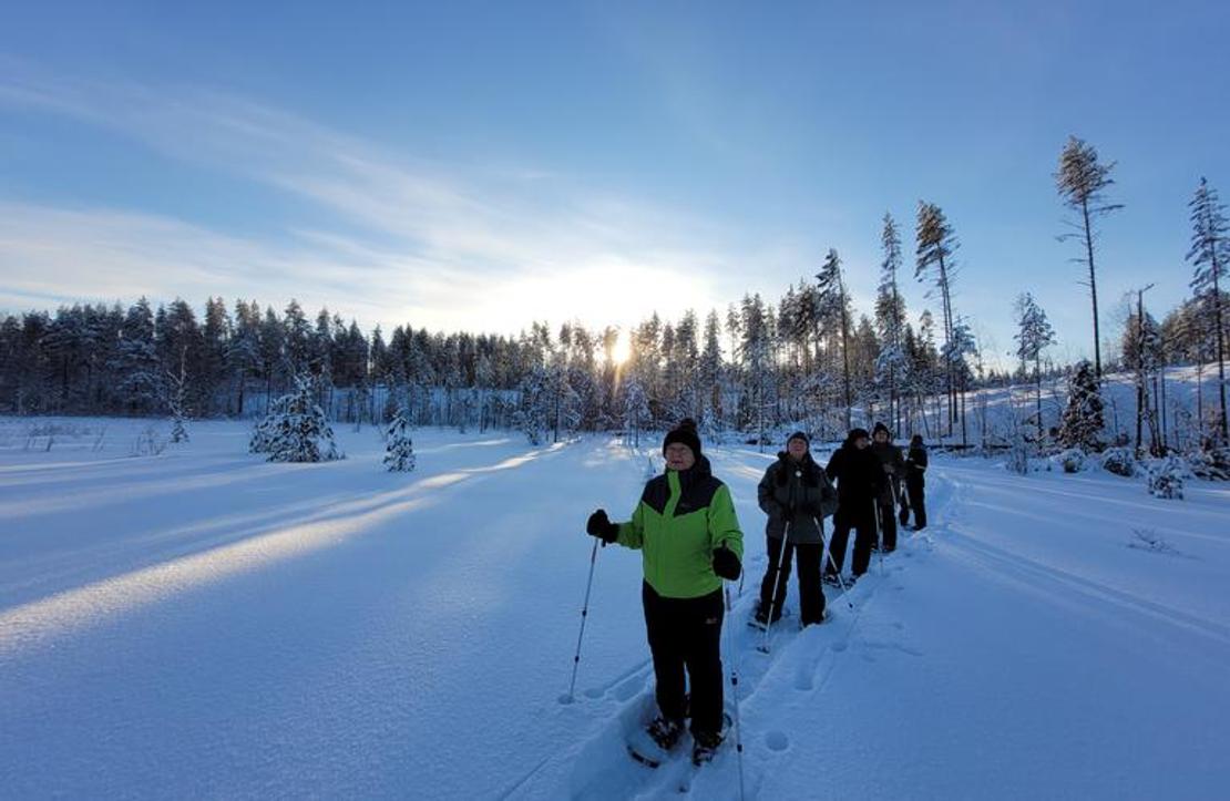 Ryhmä lumihiihdon tai lumikenkäilyn harrastajia hiihtää tai kävelee jonossa syvän, lumisen aukean yli kirkkaana talvipäivänä. Etualalla vasemmalla on yksi osallistuja kirkkaan limevihreässä takissa katsoen kameraan. Taustalla näkyy puiden ääriviivoja, jotka ovat lumen peitossa, ja aurinko paistaa matalalta horisontista luoden pitkiä varjoja.