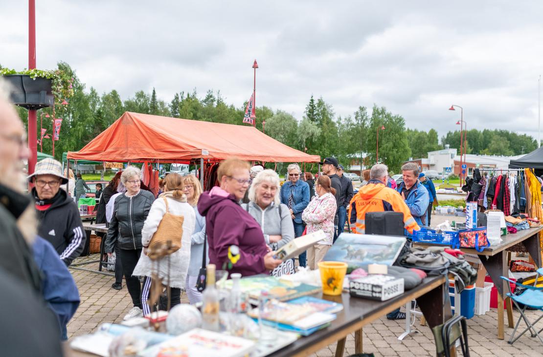 Ihmisiä tutkimassa tavaroita Ilomantsin ulkoilmatorin myyntipöydillä pilvisenä päivänä. Etualalla on pöytä täynnä pientavaraa, kuten kirjoja ja laatikoita, ja taustalla näkyy suuri oranssi myyntiteltta sekä vaaterekkejä.