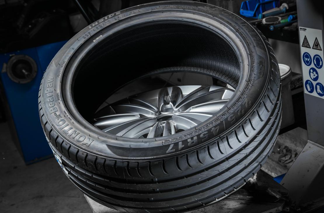 Lähikuva uudesta, matalaprofiilisesta renkaasta, joka on asennettavana kiiltävälle alumiinivanteelle. Renkaan pintakuvio on selkeä ja se on asetettu kaltevassa asennossa todennäköisesti rengaskoneen päällä. Taustalla näkyy epätarkkana sinisiä ja harmaita osia rengashuoltovälineistöä.