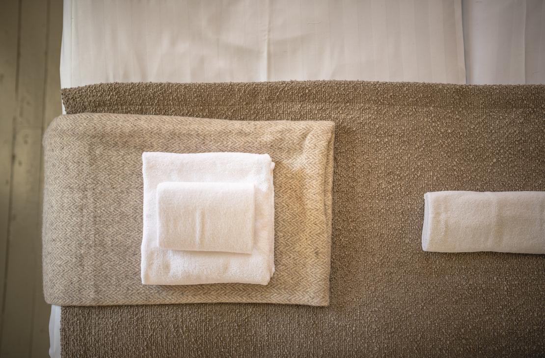 Lähikuva hotellihuoneen sängyn jalkopäästä tai päädystä Matkailukeskus Puu-Juuka Oy:ssä. Sänky on pedattu valkoisilla lakanoilla. Patjan päällä on ruskea sängynpeite tai leveä päiväpeite. Peitteen päällä on siististi taiteltu kasa valkoisia pyyhkeitä, ja oikealla on yksi erillinen pyyhe.