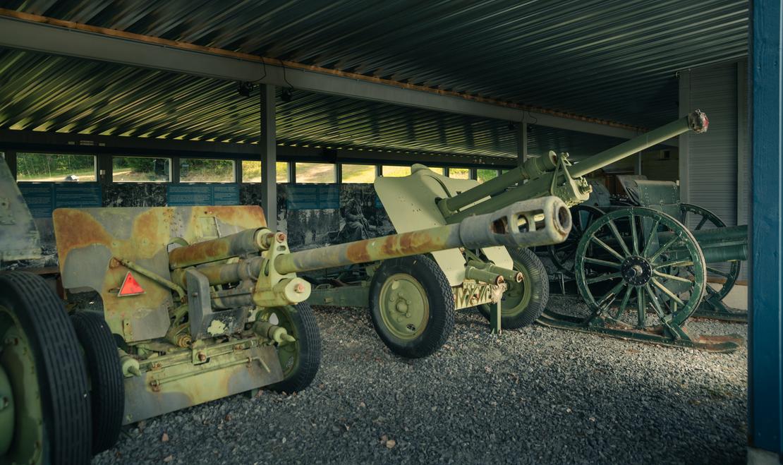Kuva Taistelijan talon tykkikatoksesta esittää rivin historiallisia tykkejä ja aseita suojassa. Etualalla on harmaa-vihreä, maastokuvioitu panssarintorjuntatykki pyörillä. Sen takana on toinen, samankaltainen tykki, ja oikealla näkyy vanhempi, vihreä kenttätykki suurilla puupyörillä. Katoksen lattia on soraa tai hiekkaa, ja tausta on tumma.