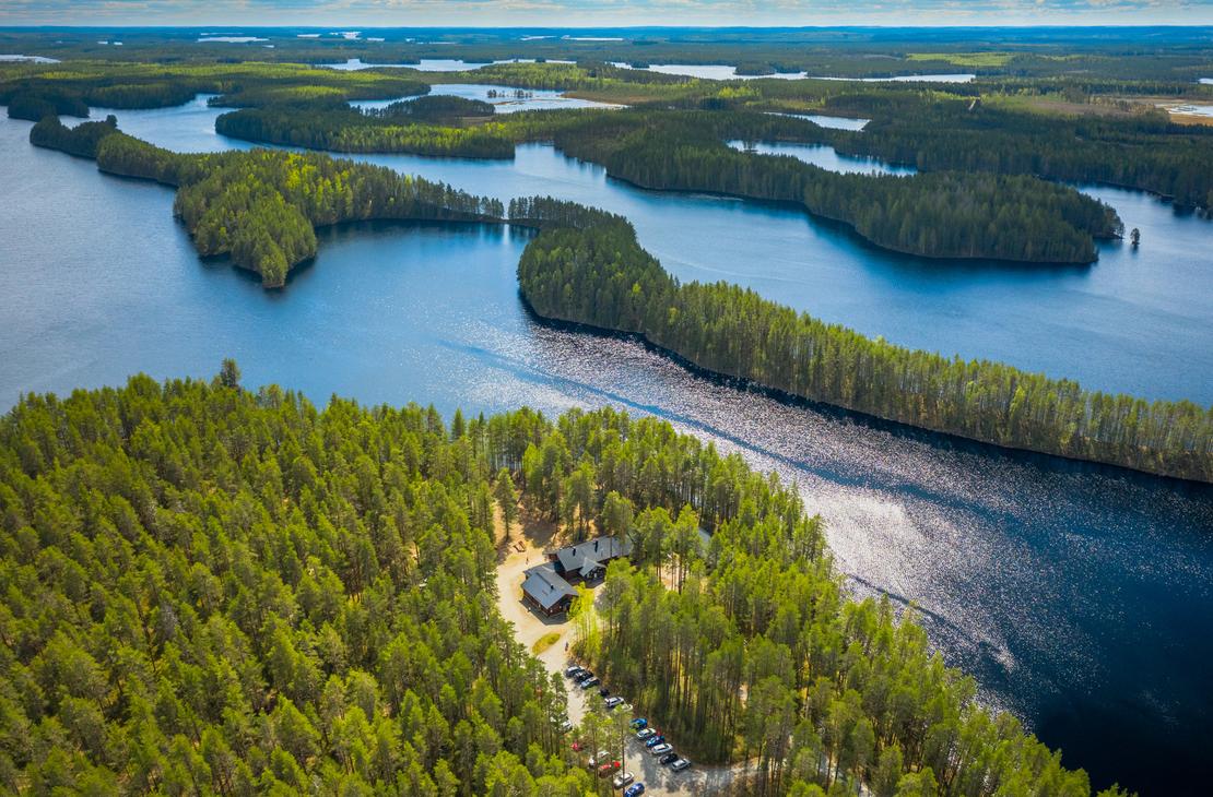 Ilmakuva Petkeljärvi Centerin alueesta ja Petkeljärven kansallispuiston maisemista. Etualalla on tiheää, vihreää metsää ja avoin, hiekkainen alue, jossa näkyy muutama tummakattoinen rakennus (Petkeljärvi Center). Taustalla avautuu laaja järvimaisema, jossa tummansininen vesi kietoutuu lukuisten vihreiden harjujen ja niemien ympärille. Kirkas sininen taivas, jossa on valkoisia pilviä.