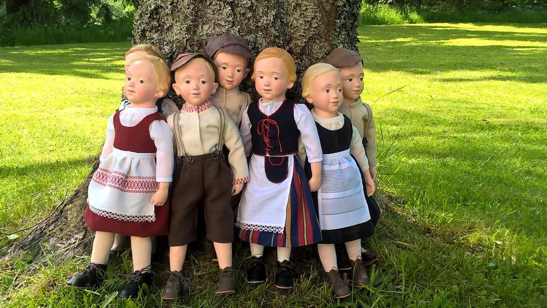 Kuva esittää kahta pehmolelua: vasemmalla on suuri, vaaleanruskea, perinteisen näköinen nallekarhu, jolla on yllään valkoinen paita ja siniset housut. Oikealla on pienempi nukke, jolla on punaiset hiukset ja yllään raidallinen paita ja tumma hame. Molemmat lelut istuvat pöydällä tai hyllyllä, ja tausta on epätarkka. Kuva on todennäköisesti peräisin Nukkemuseosta tai leluliikkeestä.