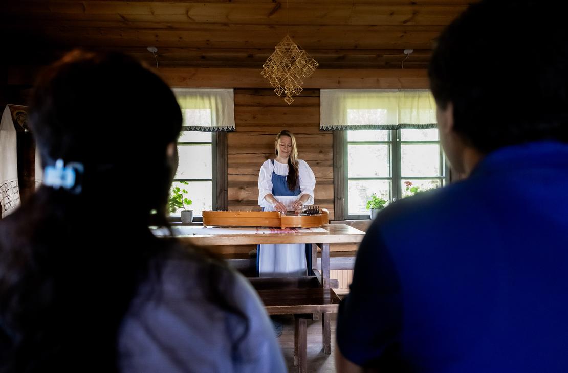 Perinteiseen asuun pukeutunut nainen soittaa kanteletta hirsituvassa, kuvattuna kahden etualalla istuvan kuuntelijan olkap&auml;iden v&auml;list&auml;. Katossa on olkihimmeli ja taustalla ikkunoita, joiden laudoilla kukkii pelargoneja.
