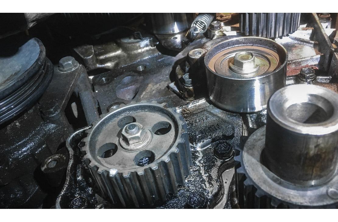 Lähikuva autonmoottorin osista, todennäköisesti jakopään alueelta. Kuvassa näkyy kaksi hammaspyörää ja yksi kiristinrulla tai vastaava metallinen pyörä. Osat ovat tummat, kiiltävät ja mahdollisesti öljyiset. Kohtaus on otettu moottoritilassa korjauksen tai huollon aikana.