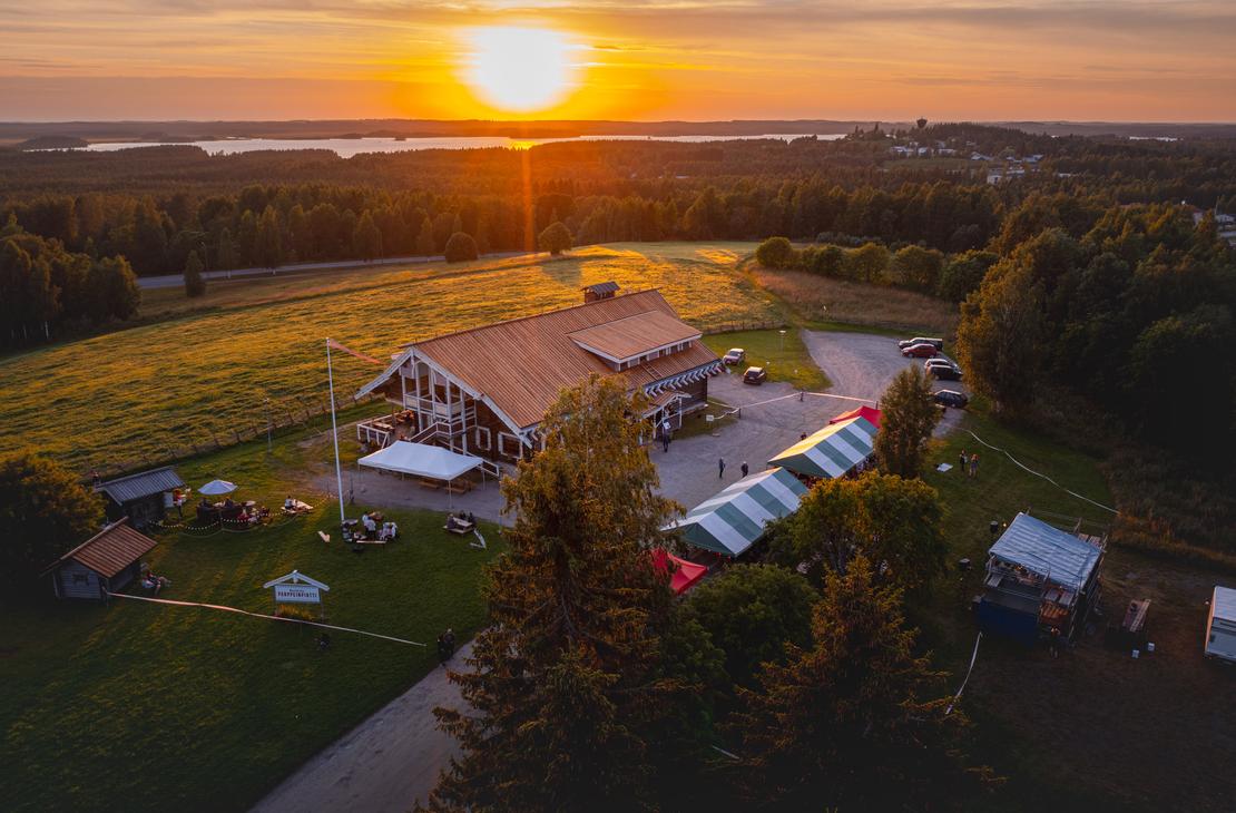 Ilmakuva Parppeinpirtistä ja sitä ympäröivästä metsä- ja järvimaisemasta kirkkaassa auringonlaskussa. Laskeva aurinko heijastaa kultaista valoa koko näkymään.