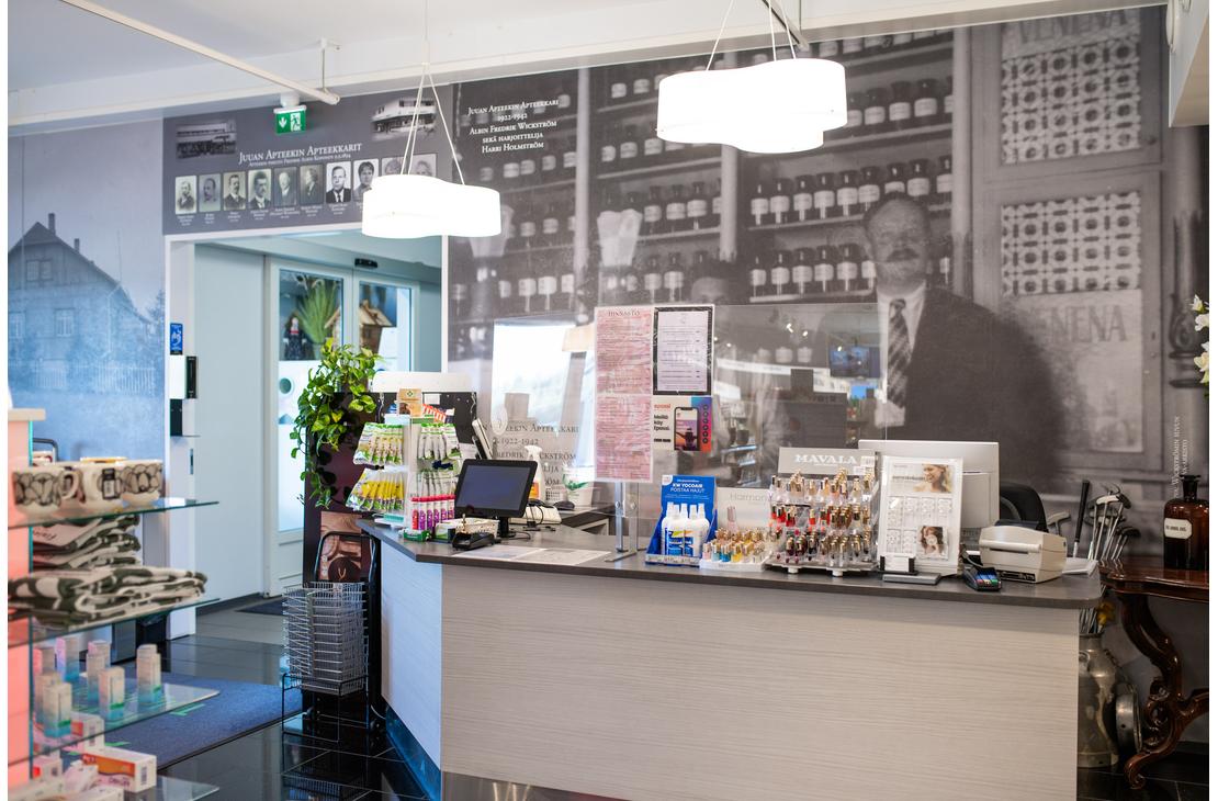 Kuva esittää Juuan Apteekin sisätilaa ja palvelutiskiä. Etualalla on harmaa palvelutiski, jonka takana on seinällä mustavalkoinen, suuri valokuva vanhanaikaisesta apteekista tai apteekkarista hyllyineen. Tiskillä on erilaisia apteekkituotteita. Tila on hyvin valaistu.