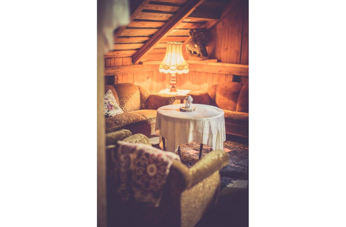 Kuva esittää Pappila Marjamäen ullakolla tai parvella sijaitsevaa oleskelutilaa. Tila on sisustettu kodikkaasti retrotyylisillä, sammaleenvihreillä tai oranssinruskeilla sohvilla ja nojatuoleilla. Huoneessa on puupaneeliseinät ja -katto. Keskellä on pyöreä pöytä valkoisella pöytäliinalla, ja tila on valaistu lämpimästi vanhanaikaisella pöytävalaisimella. Taustalla näkyy kattorakenteita ja ylätasolle vievät tikkaat.