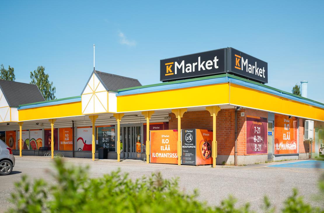 K-Market Ilomantsi -ruokakaupan julkisivu ja sis&auml;&auml;nk&auml;ynti aurinkoisena p&auml;iv&auml;n&auml; sinisen taivaan alla. Tiilirakennuksen katolla on suuret mustat K-Market-kyltit, ja katon reunaa kiert&auml;&auml; keltainen lippa. Ikkunoissa ja seiniss&auml; on oransseja opasteita, joissa lukee &rdquo;ILO EL&Auml;&Auml; ILOMANTSISSA&rdquo; sek&auml; kaupan aukioloajat. Sis&auml;&auml;nk&auml;ynnin l&auml;hell&auml; on noutopistekaappi, ja etualalla kasvaa vihre&auml;&auml; pensaikkoa.